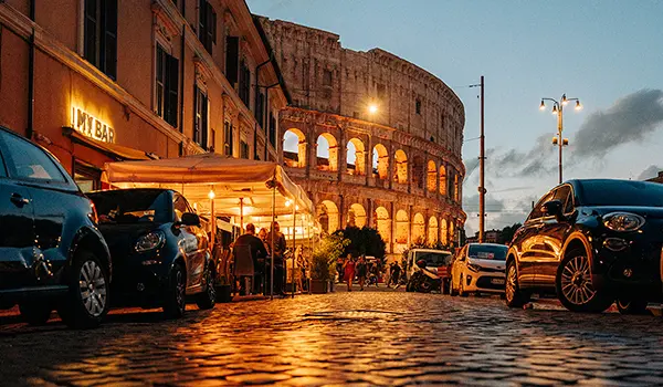 Photographie urbaine nocturne par Speetch : vue sur le Colisée illuminé à Rome depuis une rue pavée animée, démontrant une expertise en gestion des lumières de nuit.